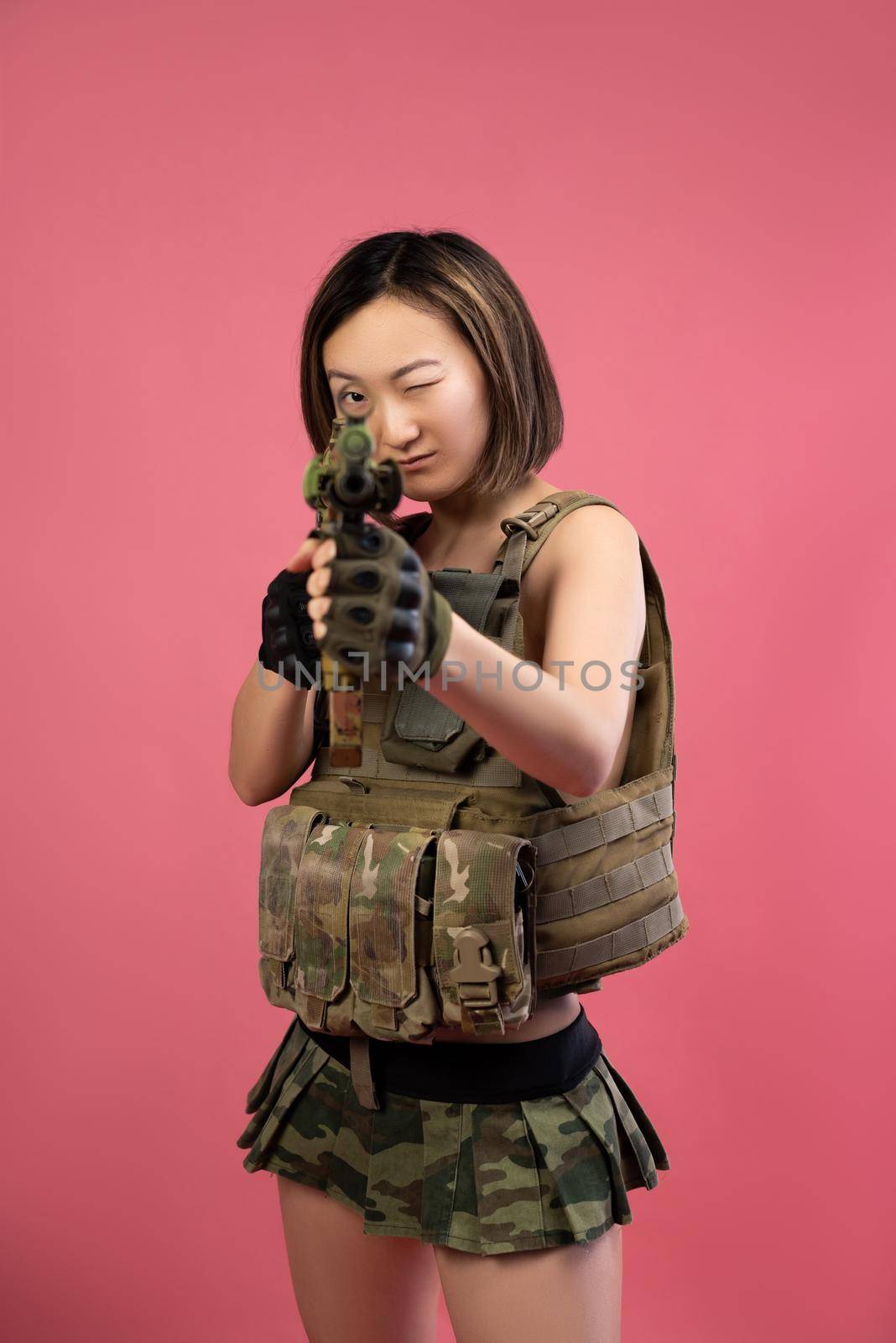 sexy Asian woman in military clothes with an automatic rifle in her hands on a pink background by Rotozey