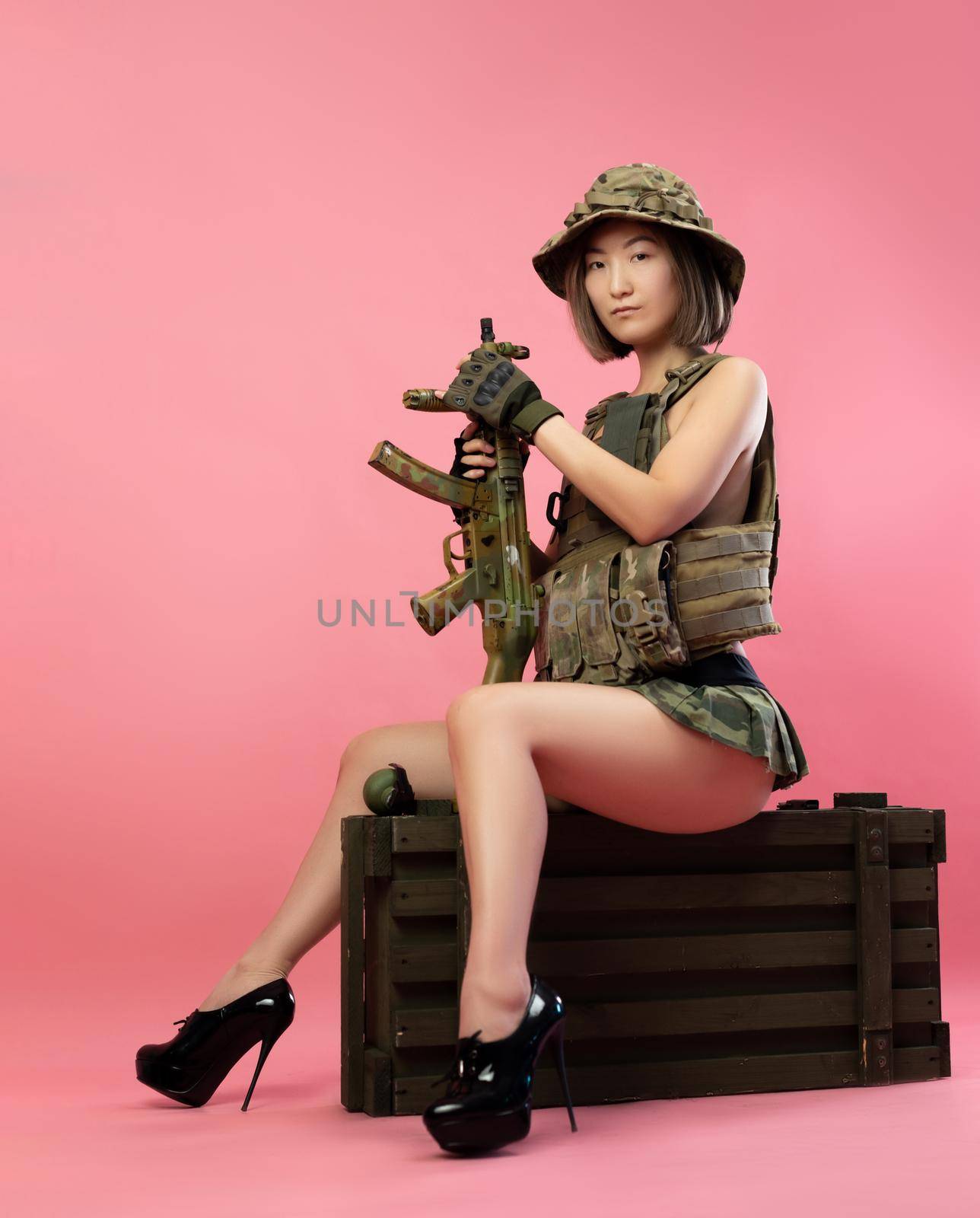 sexy Asian woman in military uniform with an automatic rifle sits on an ammunition crate by Rotozey