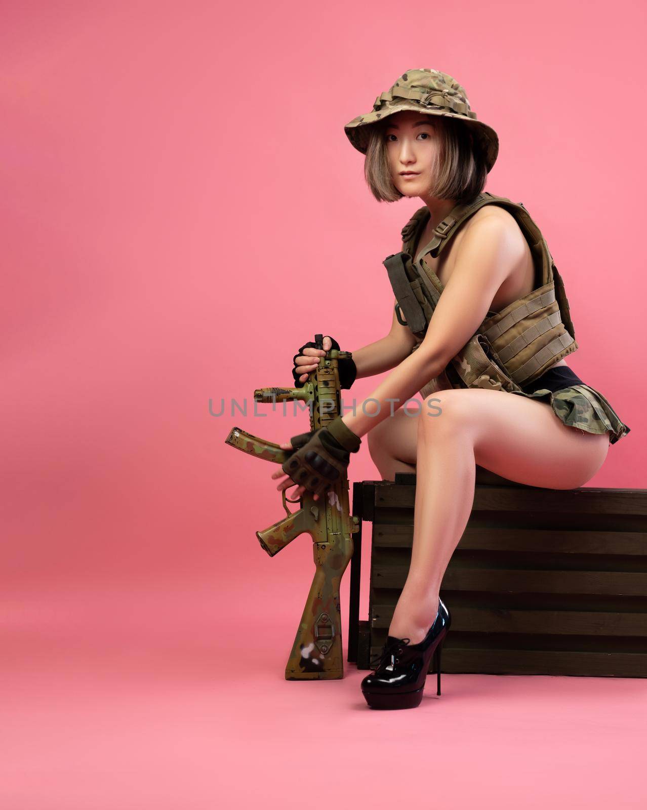 the sexy Asian woman in military uniform with an automatic rifle sits on an ammunition crate