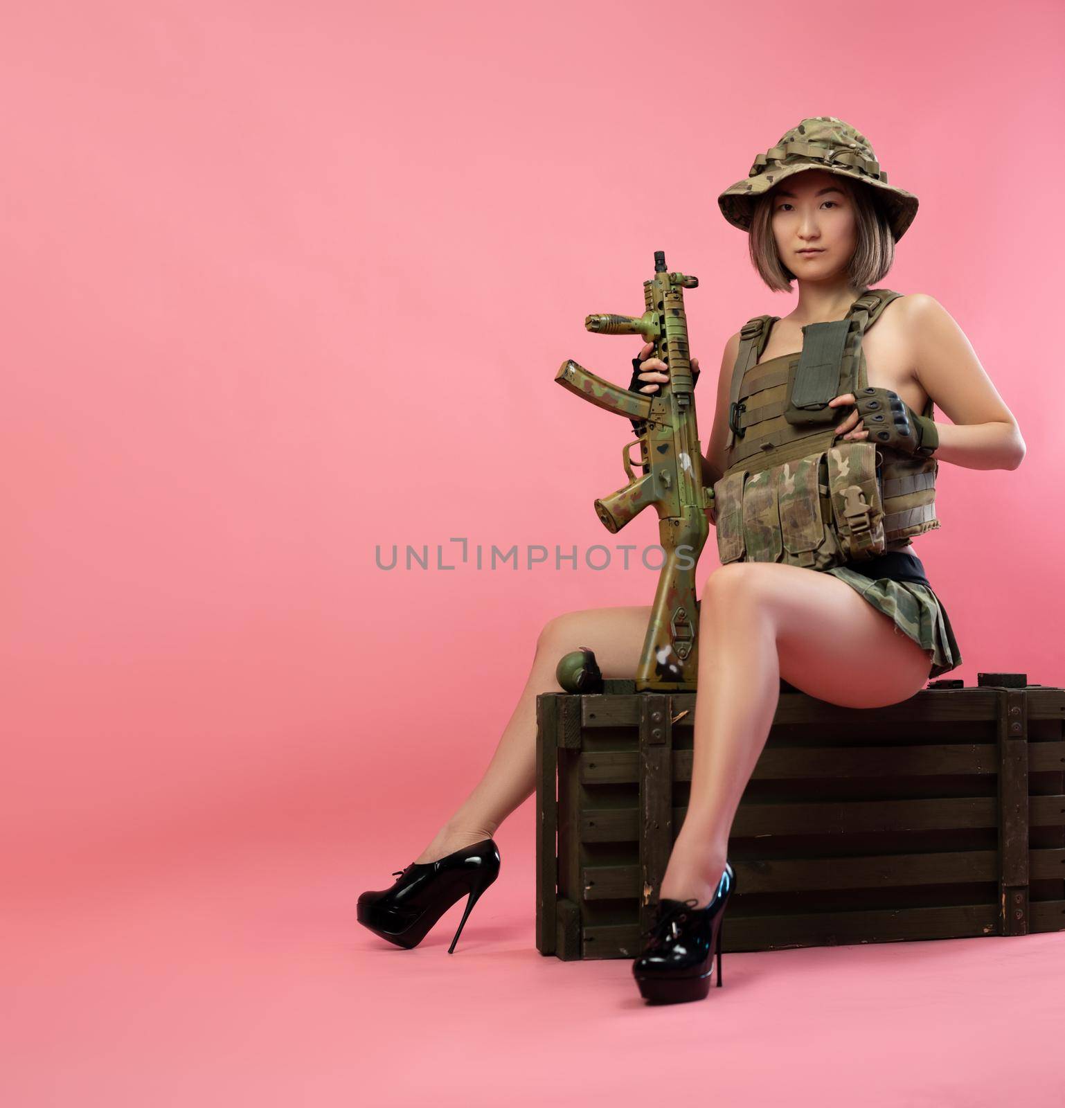 the sexy Asian woman in military uniform with an automatic rifle sits on an ammunition crate