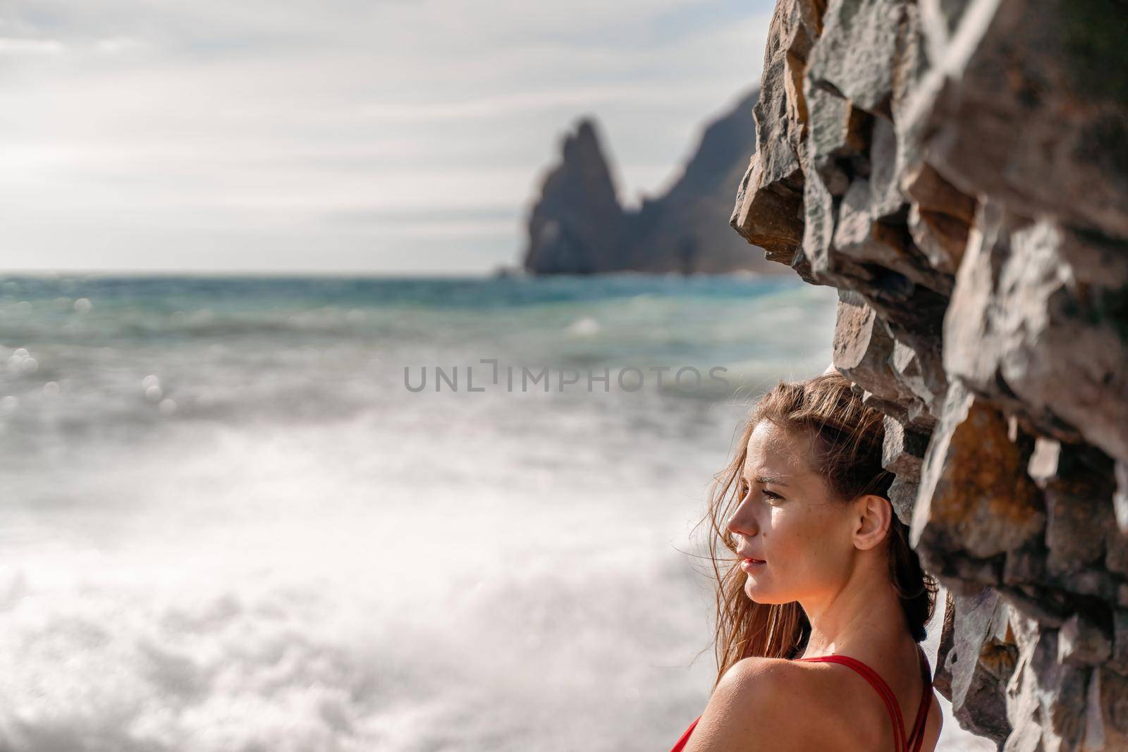 A beauty in a red swimsuit with long legs poses on a fantastic beach with huge waves against the background of mountains by Matiunina