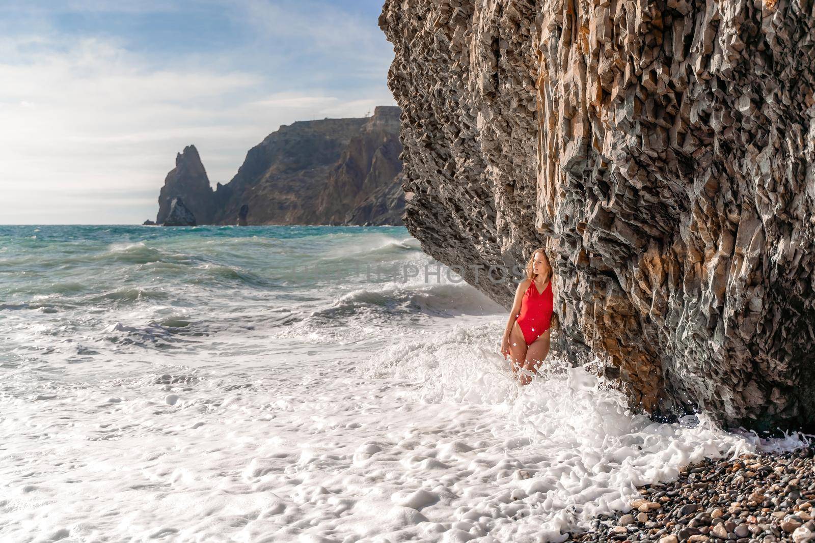 A beauty in a red swimsuit with long legs poses on a fantastic beach with huge waves against the background of mountains.