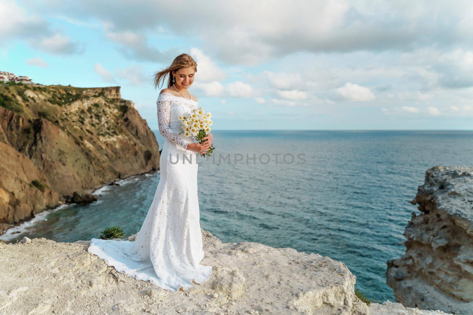Summer bouquet of field daisies in the hands of a bride in a white dress against the backdrop of the sea. She stands on a rock above the sea. Copy space. The concept of calmness, silence and unity with nature