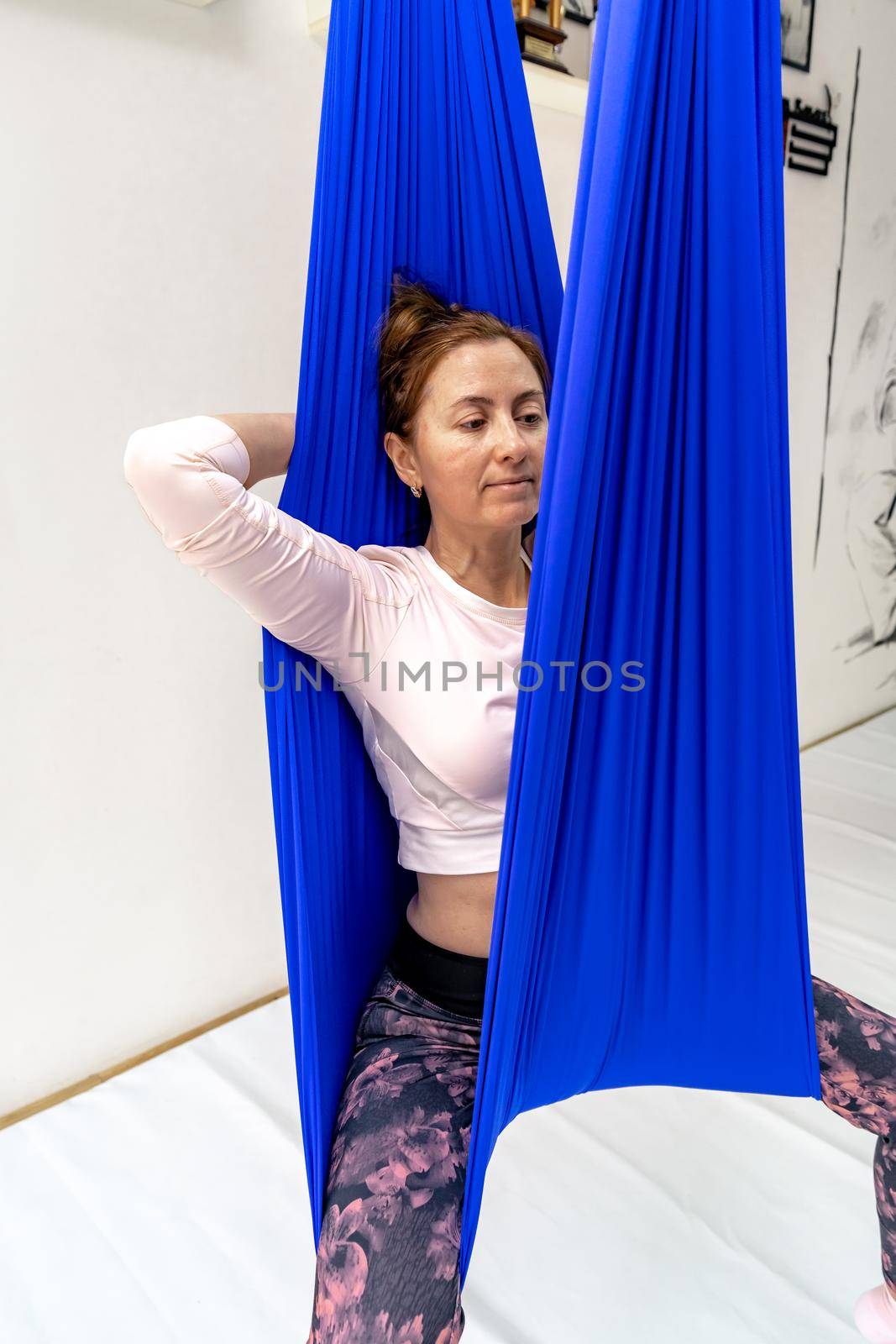 A young woman poses while doing anti-gravity aerial yoga in a blue hammock on a white background