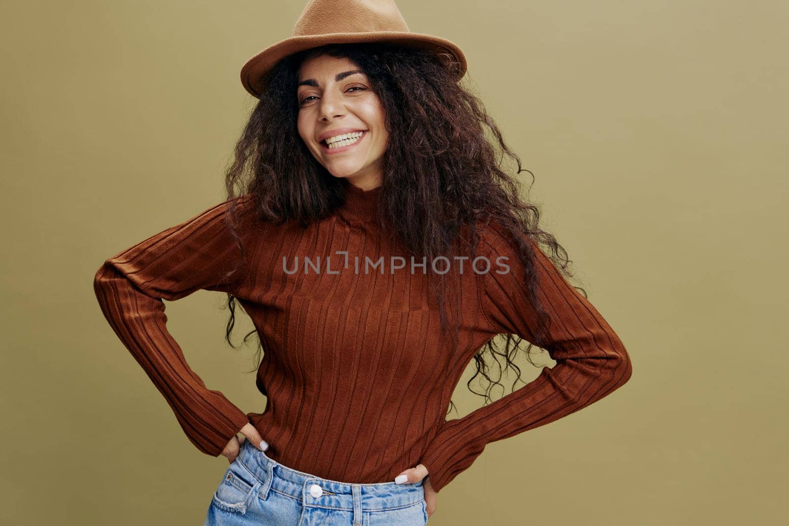 Enjoyed stylish curly Latin female in brown hat, smiling at camera, holding hand on waist, saying Yeah, isolated green background. Copy space clothing fashion brands, free place for your ad.