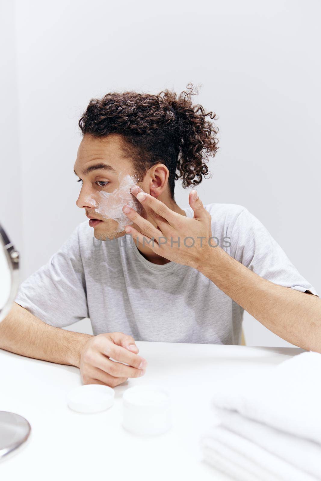 A young man in a white T-shirt sits at a table with a mirror and a towel cream on his face isolated background by SHOTPRIME