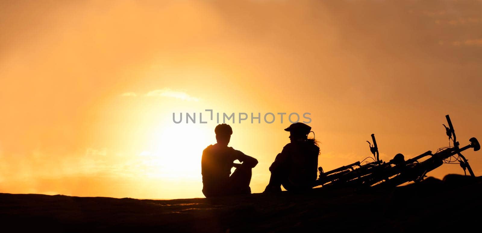 Silhouette of a couple watching the sunset with their bicycles beside them at the beach.