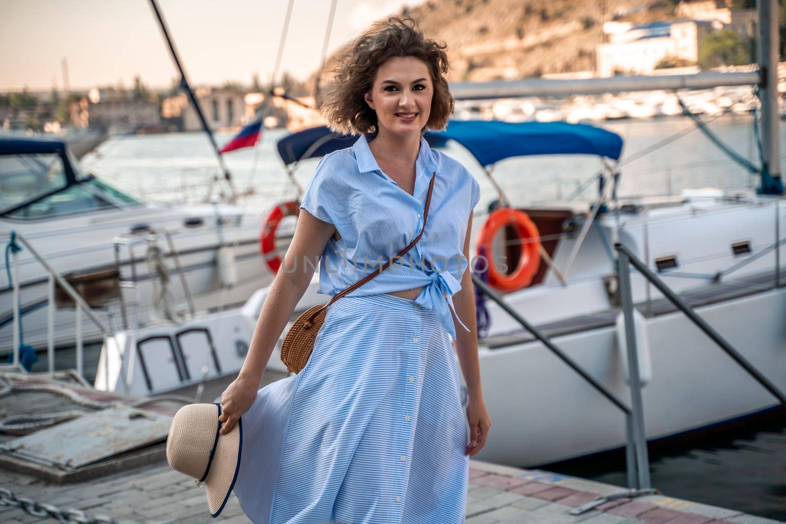 A young happy woman in a blue dress and hat stands near the seaport with luxury yachts. Travel and vacation concept.