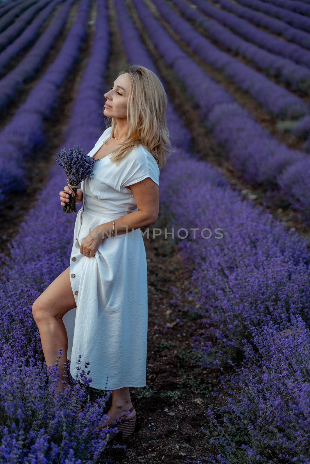 A middle-aged woman, blonde in a white dress, stands in a lavender field with a bouquet of lavender. A large flowering field in full bloom