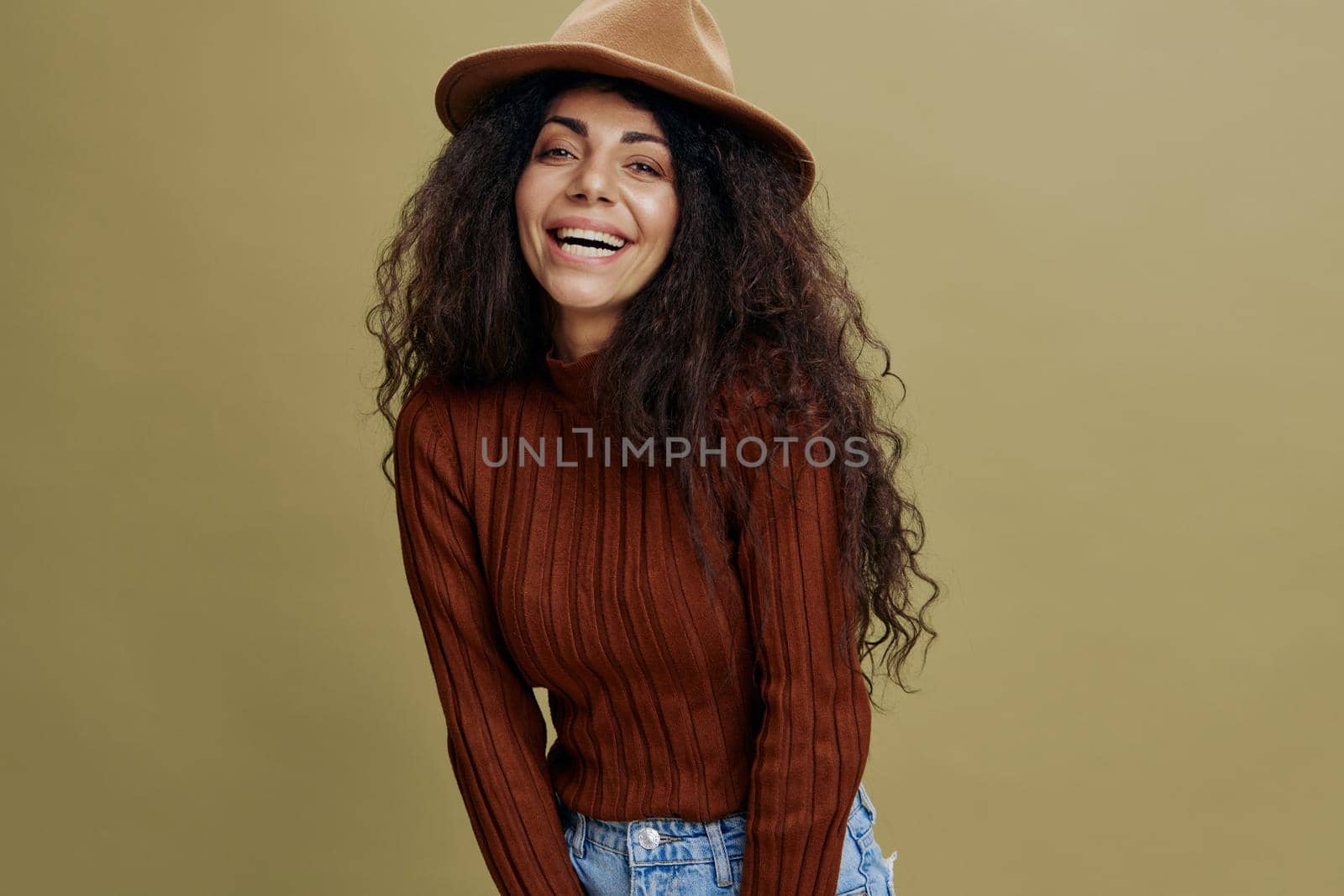 Enjoyed stylish curly Latin female in brown hat, smiling at camera, holding hand on hips, saying Yeah, isolated green background. Copy space clothing fashion brands, free place for your ad. by SHOTPRIME