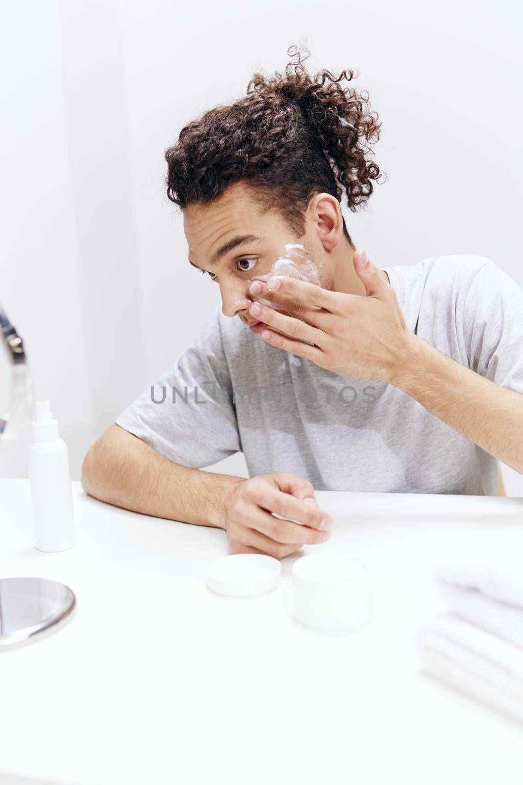 guy with curly hair puts cream on the face but sitting at the table in front of the mirror isolated background by SHOTPRIME