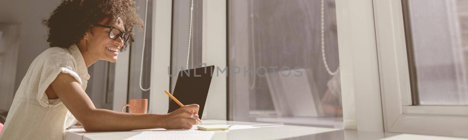 Side view of happy African American woman sitting in coworking near the window while using laptop