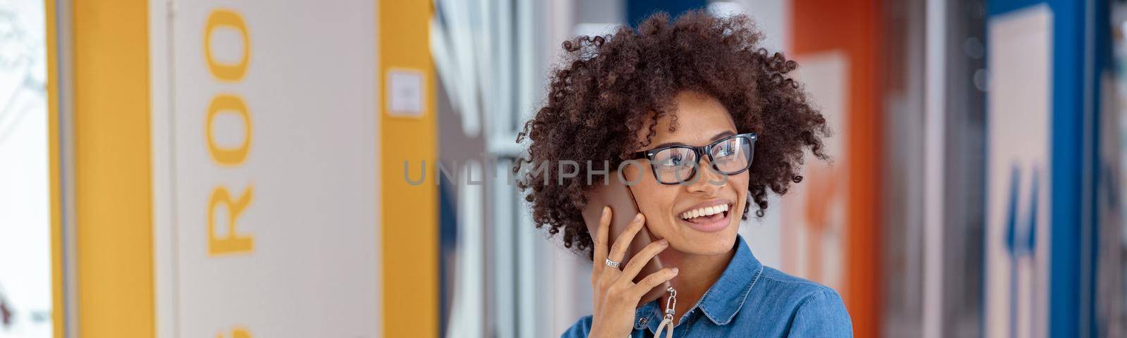 Portrait of smiling Afro American woman in glasses standing in office corridor while talking on smartphone