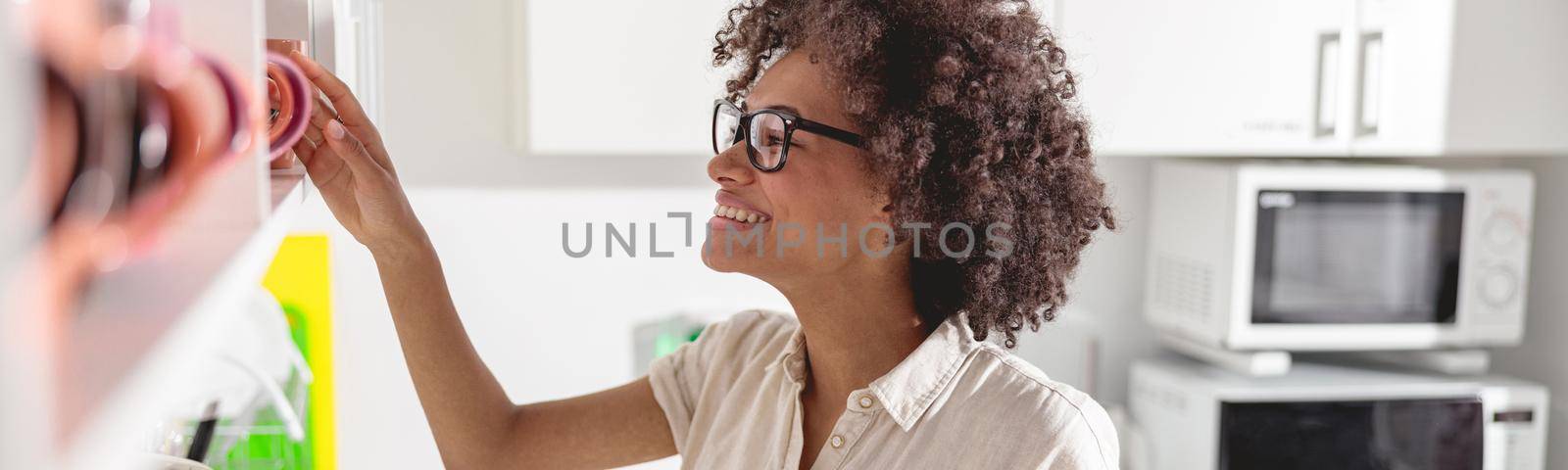 Smiling young woman holding smartphone while going to make coffee on office kitchen