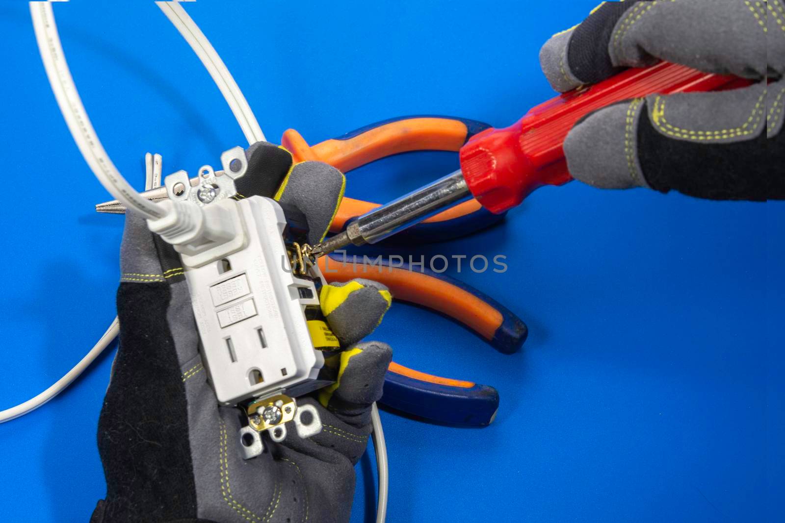 An electrician holds a power socket with a hand in a glove and fixes a screw with a screwdriver