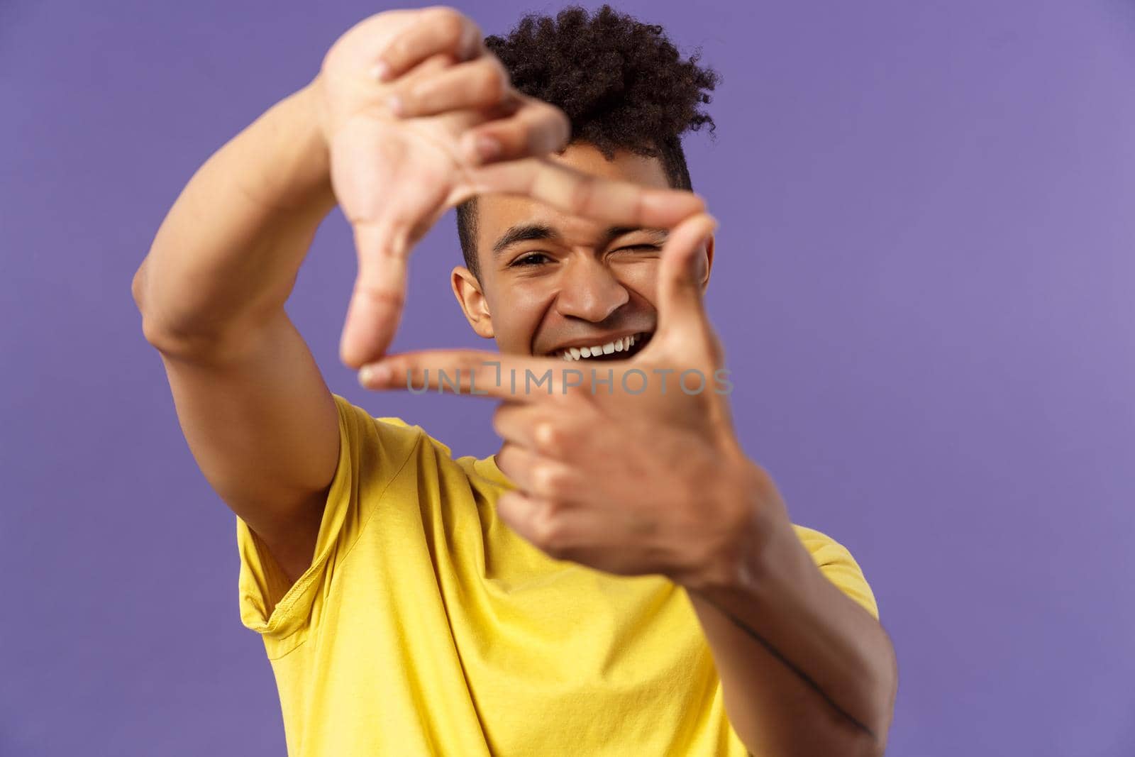 Close-up portrait of young creative photographer, man imaging, searching right angle, looking through photo frame, wink and smiling pleased, standing purple background found good shot.