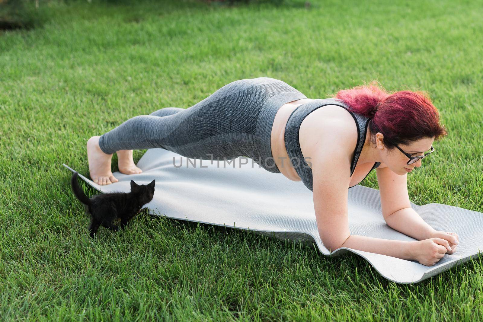 Young plus size woman in sporty top and leggings standing in plank on yoga mat spending time on green grass in yard. Black kitten walks around her. Well being and fitness concept by Satura86
