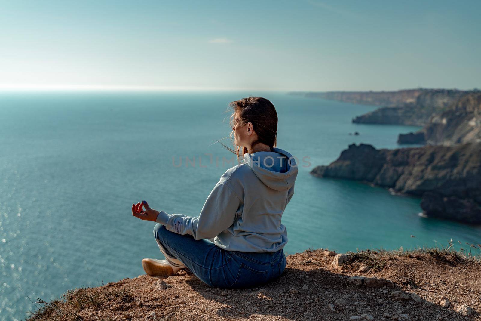 Woman tourist enjoying the sunset over the sea mountain landscape. Sits outdoors on a rock above the sea. She is wearing jeans and a blue hoodie. Healthy lifestyle, harmony and meditation.