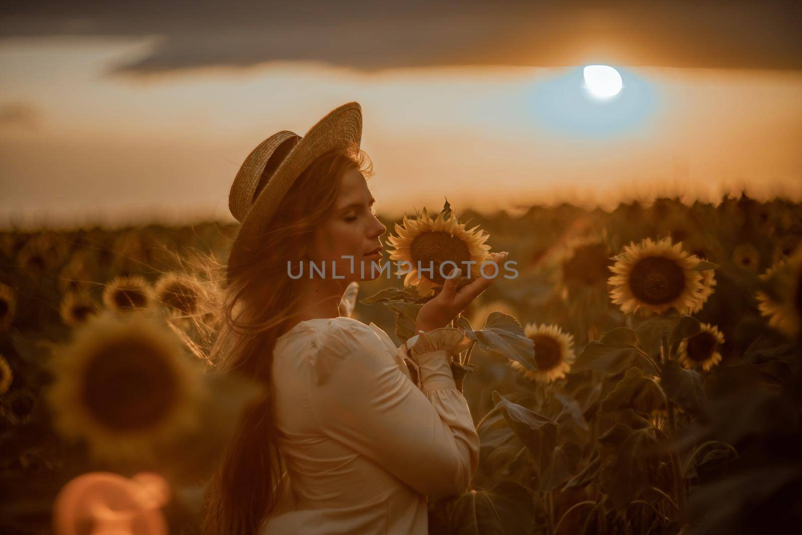 Beautiful young woman in a hat enjoying nature on a field of sunflowers at sunset. Summer. Attractive brunette with long healthy hair. by Matiunina