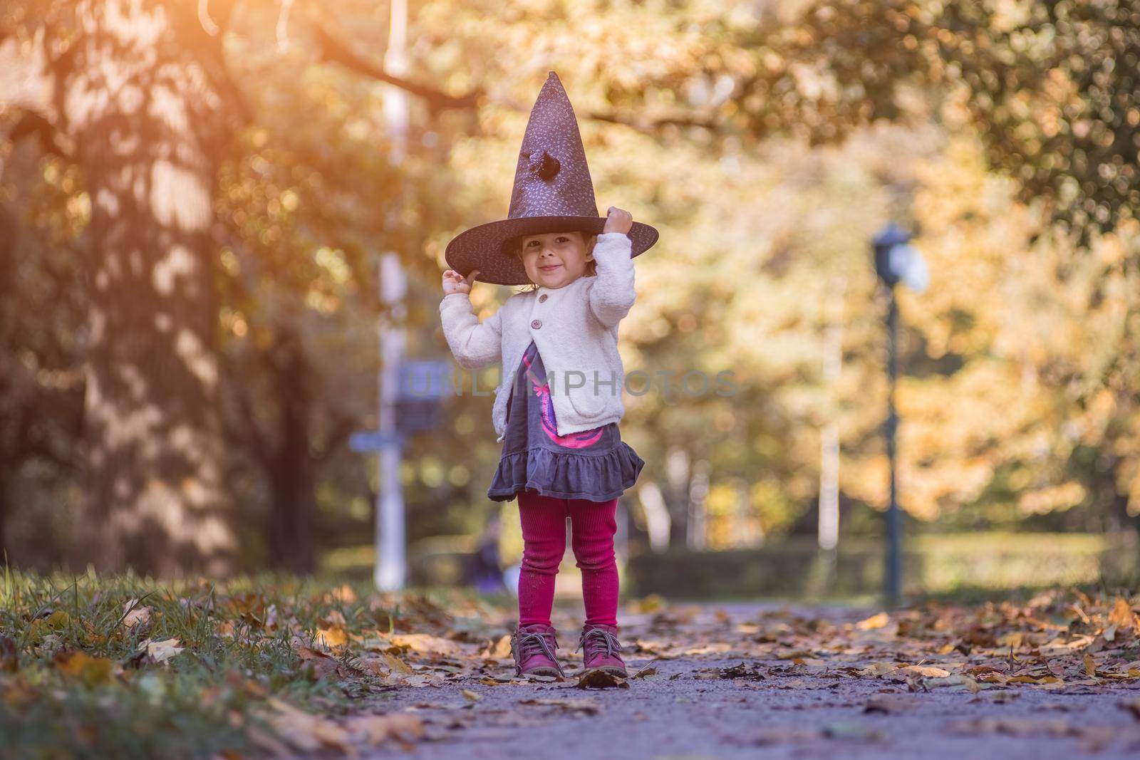 HALLOWEEN CELEBRATION CARD. Cute toddler girl in black witch hat holds her hands and smiles in autumn forest on sunny day.