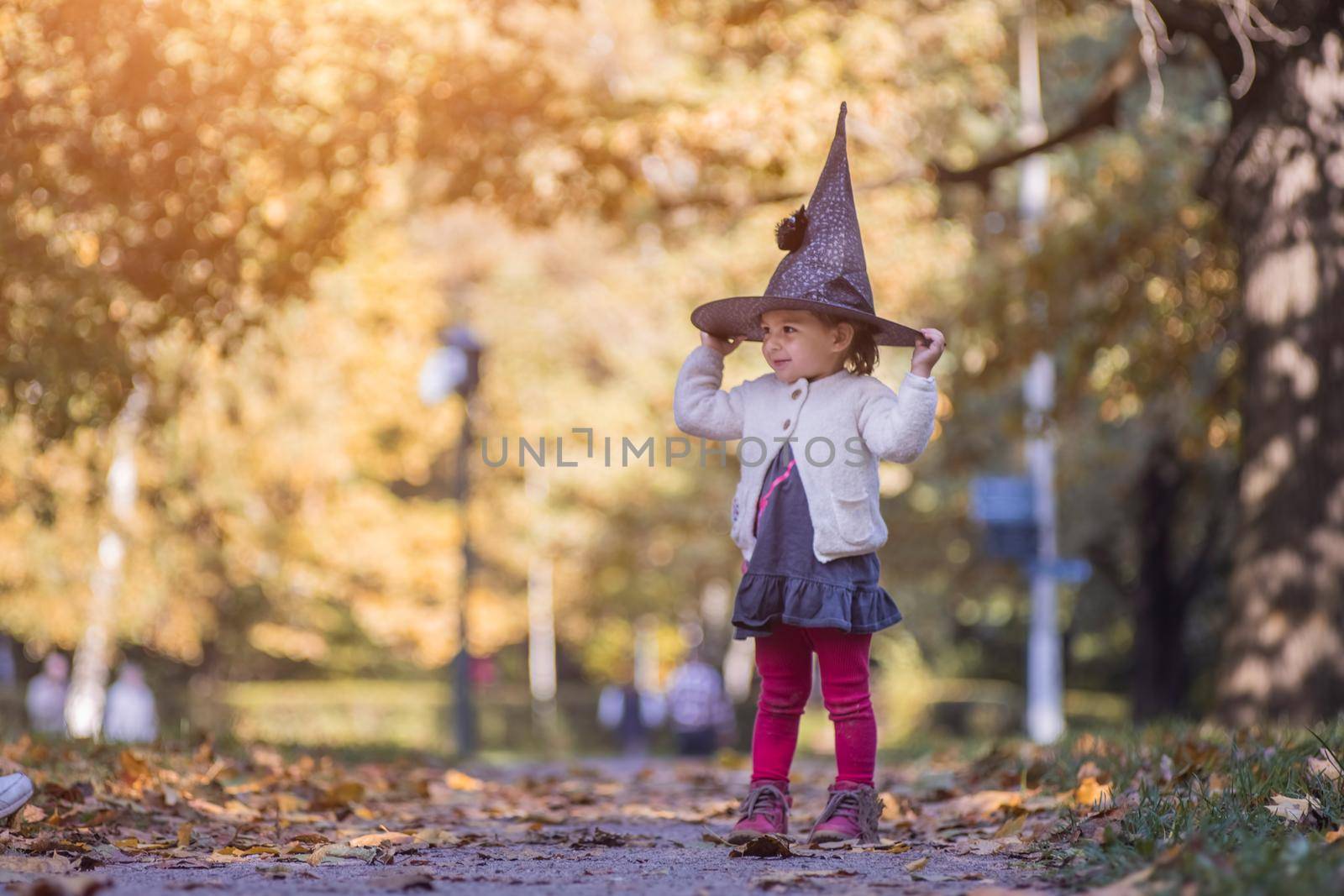 HALLOWEEN CELEBRATION CARD. Cute toddler girl in black witch hat holds her hands and smiles in autumn forest on sunny day