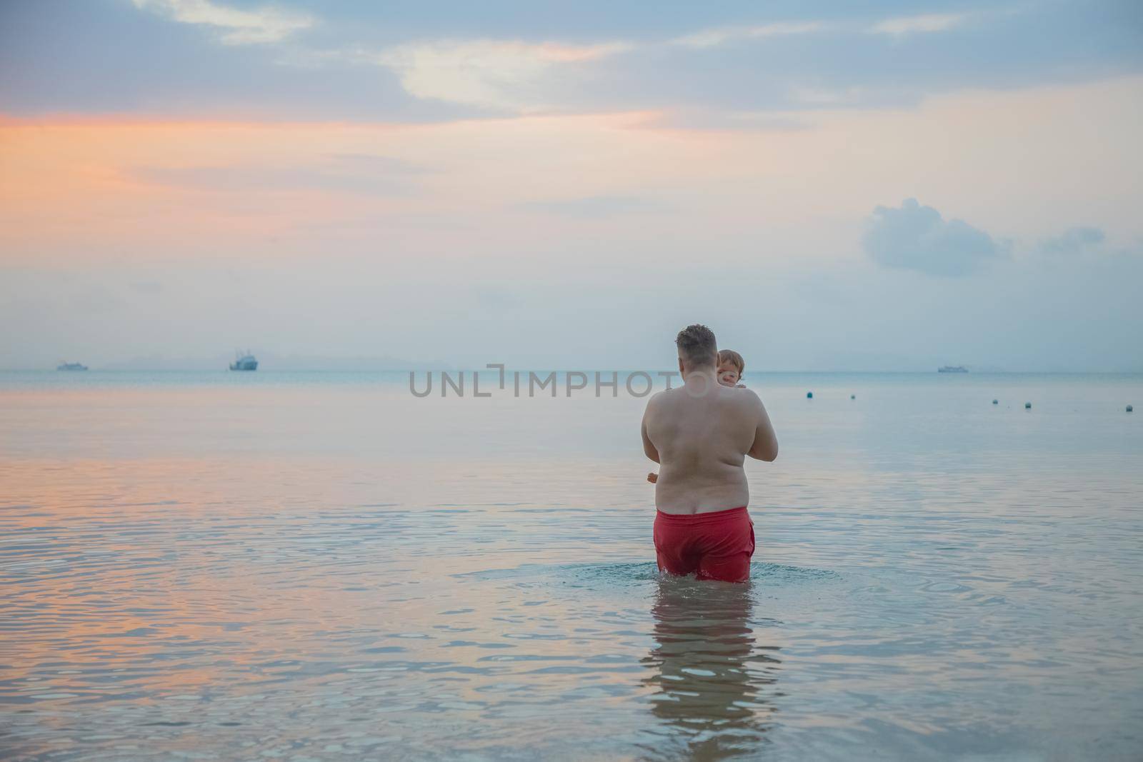 father with little child in hands enter sea water sunset time rear view. people from behind