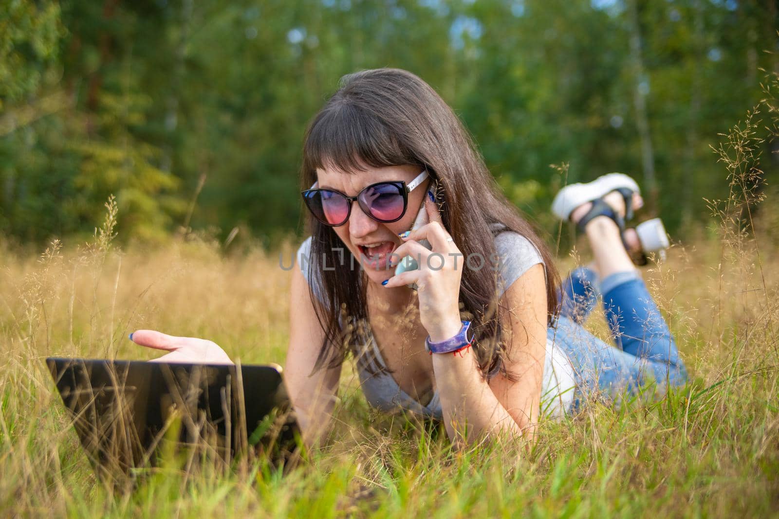 young woman swears on the phone while looking into a laptop. woman freelancer works in a sunny meadow