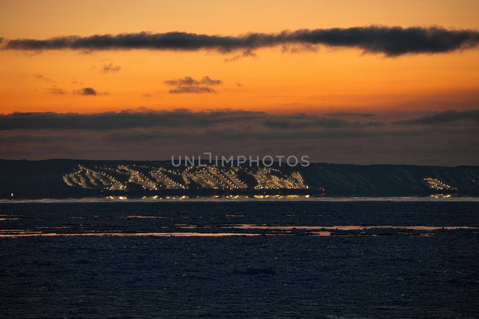 Long telephoto View of Collingwood and Blue Mountain and Craigleith Ski Hills Across Georgian Bay between sunset and twilight in winter. The ski hills are lit up with night lights and the sky is a rich sunset orange color and ice pack is gathered in the bay. High quality photo
