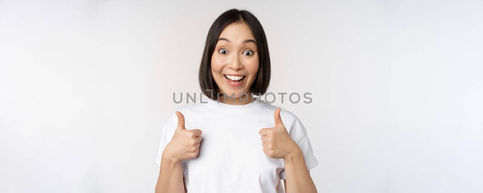 Happy young korean woman showing thumbs up and smiling, give positive feedback, recommending smth, standing over white background by Benzoix