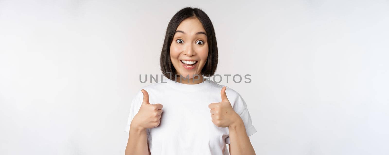 Happy young korean woman showing thumbs up and smiling, give positive feedback, recommending smth, standing over white background.