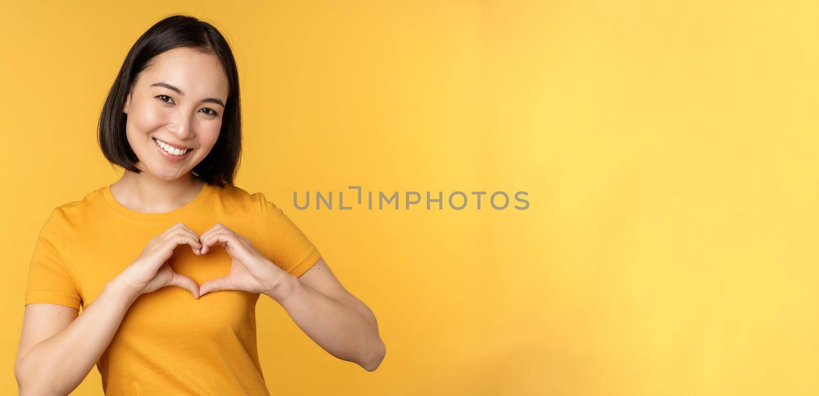 Beautiful asian girl showing heart, love gesture and smiling white teeth, express care and sympathy, standing over yellow background.
