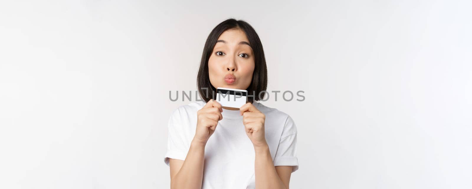 Money and finance concept. Cute japanese girl kissing her credit card, standing in tshirt over white background by Benzoix