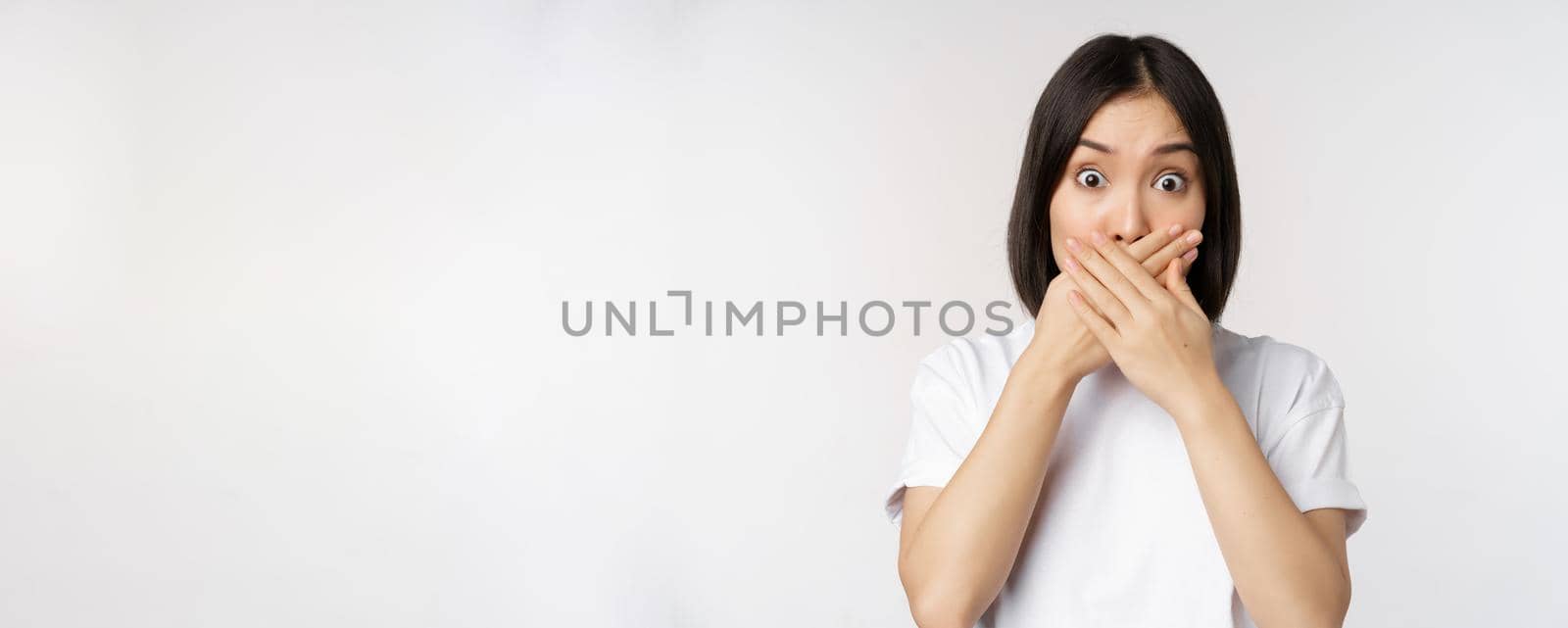 Image of shocked young asian woman shut mouth, looking startled, speechless, standing over white background by Benzoix