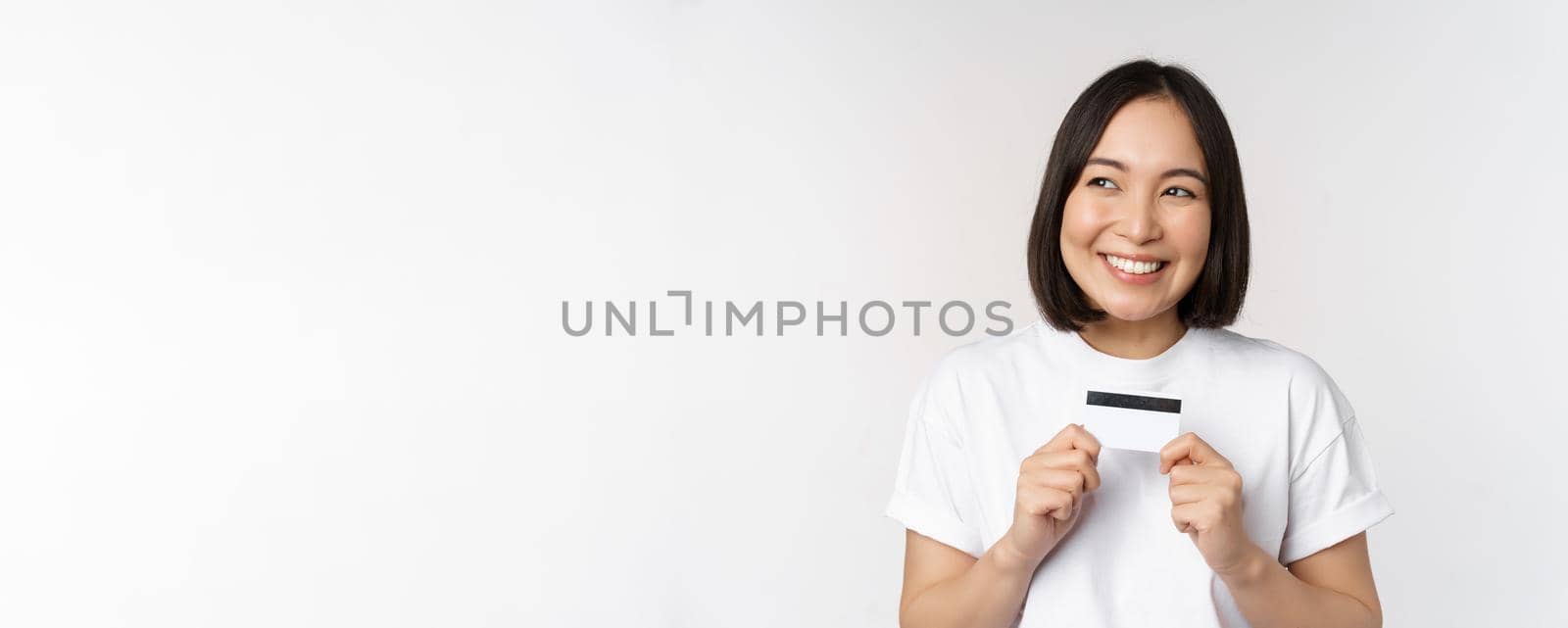 Smiling korean woman showing credit card with happy face, standing in tshirt over white background by Benzoix