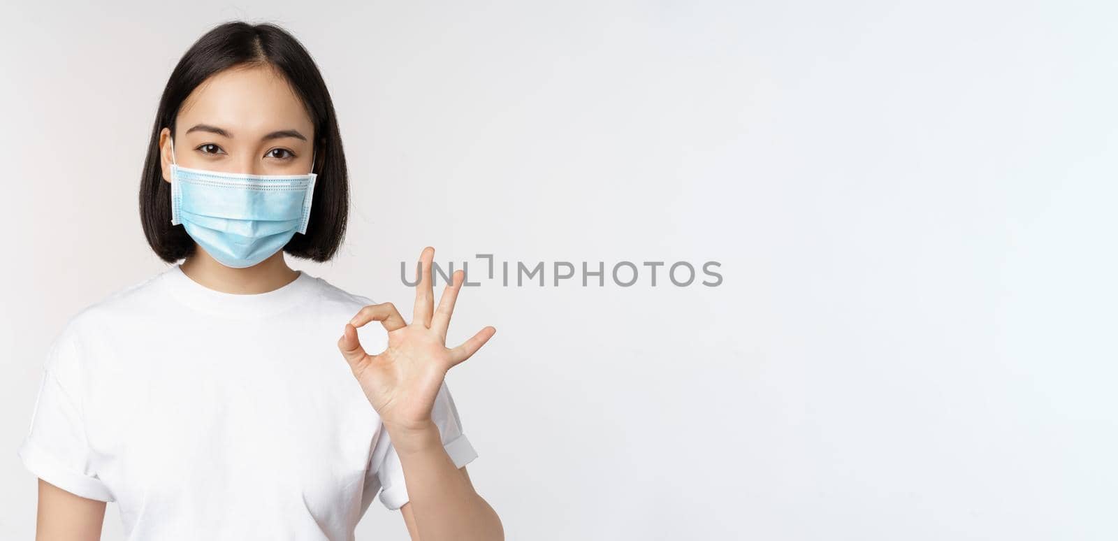 Covid-19, healthcare and medical concept. Happy asian girl smiling, wearing medical face mask, showing okay, ok sign, approve, recommending smth, white background by Benzoix