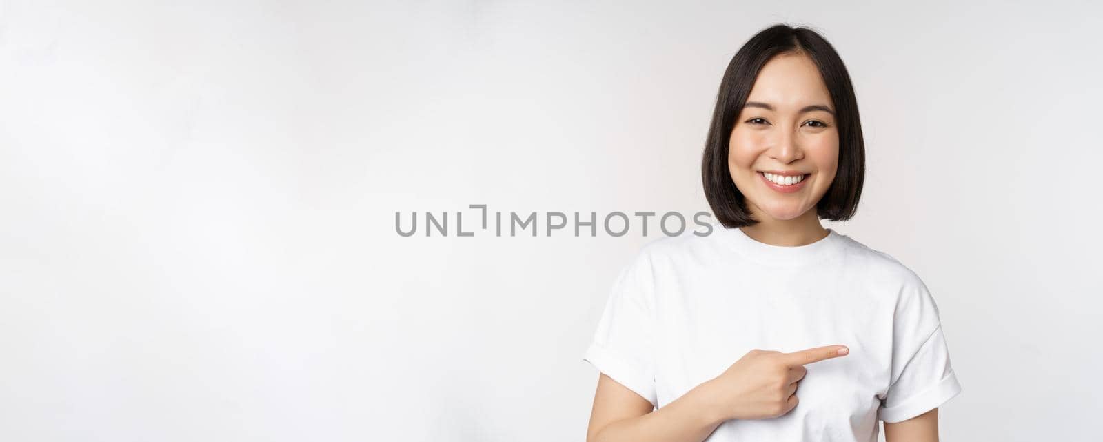 Happy beautiful asian girl showing advertisement on empty copy space, pointing finger right and smiling, demonstrating banner or company logo, white background.