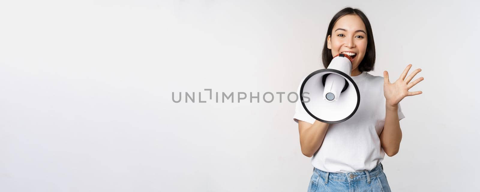 Happy asian woman shouting at megaphone, making announcement, advertising something, standing over white background.