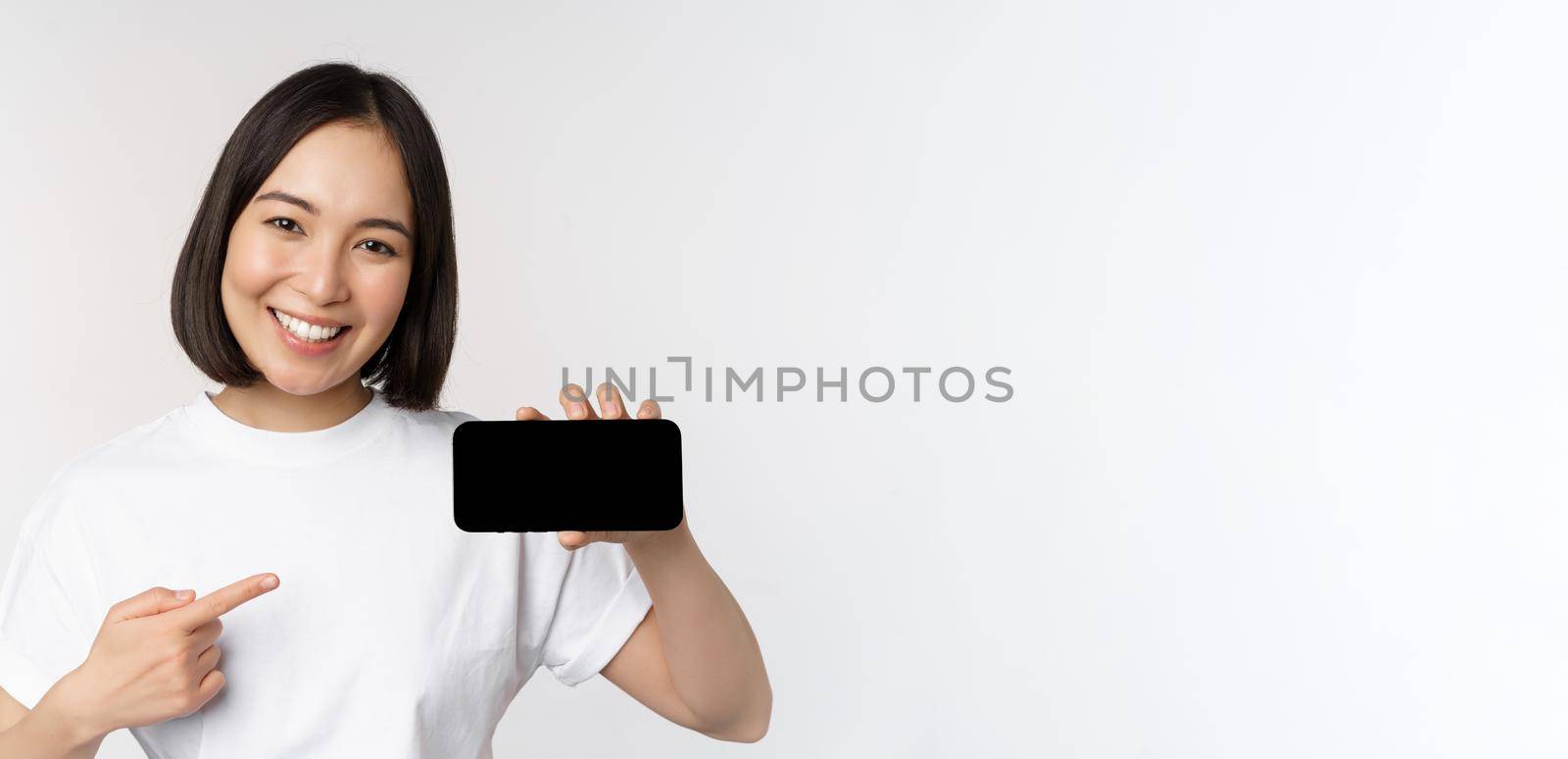 Portrait of smiling korean woman pointing finger at mobile phone screen, showing horizontal smartphone display, recommending website or store online, white background.
