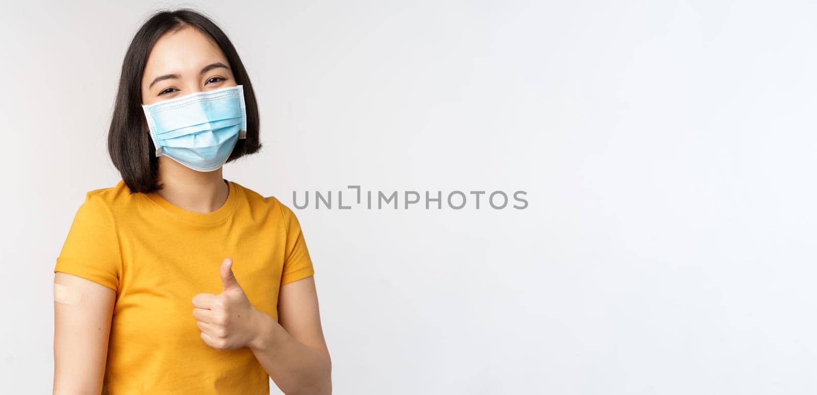 Covid-19, vaccination and healthcare concept. Portrait of cute asian girl in medical mask, has band aid on shoulder after coronavirus vaccine, standing over white background by Benzoix