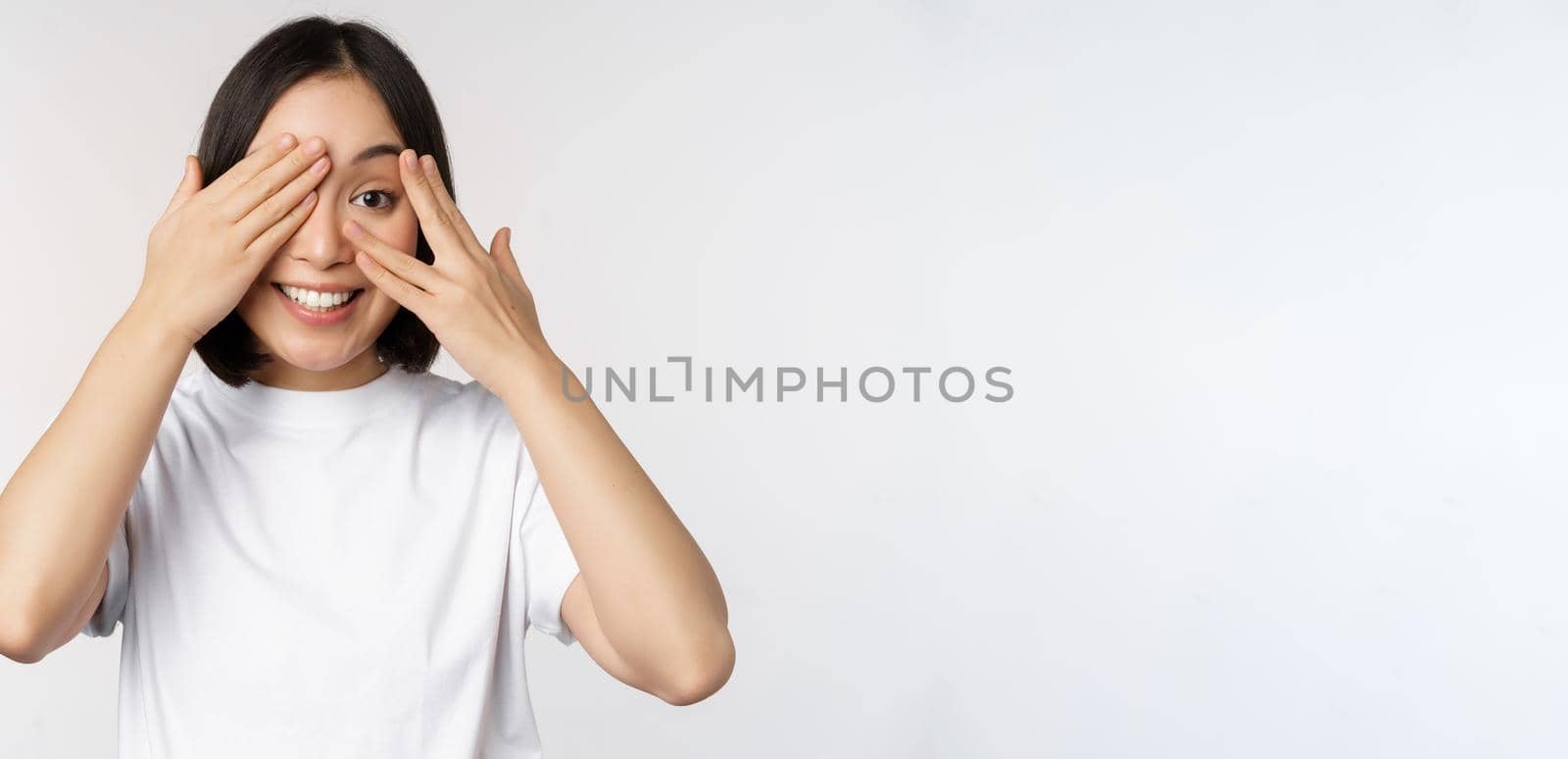 Portrait of asian woman covering eyes, waiting for surprise blindfolded, smiling and peeking at camera, standing over white background by Benzoix