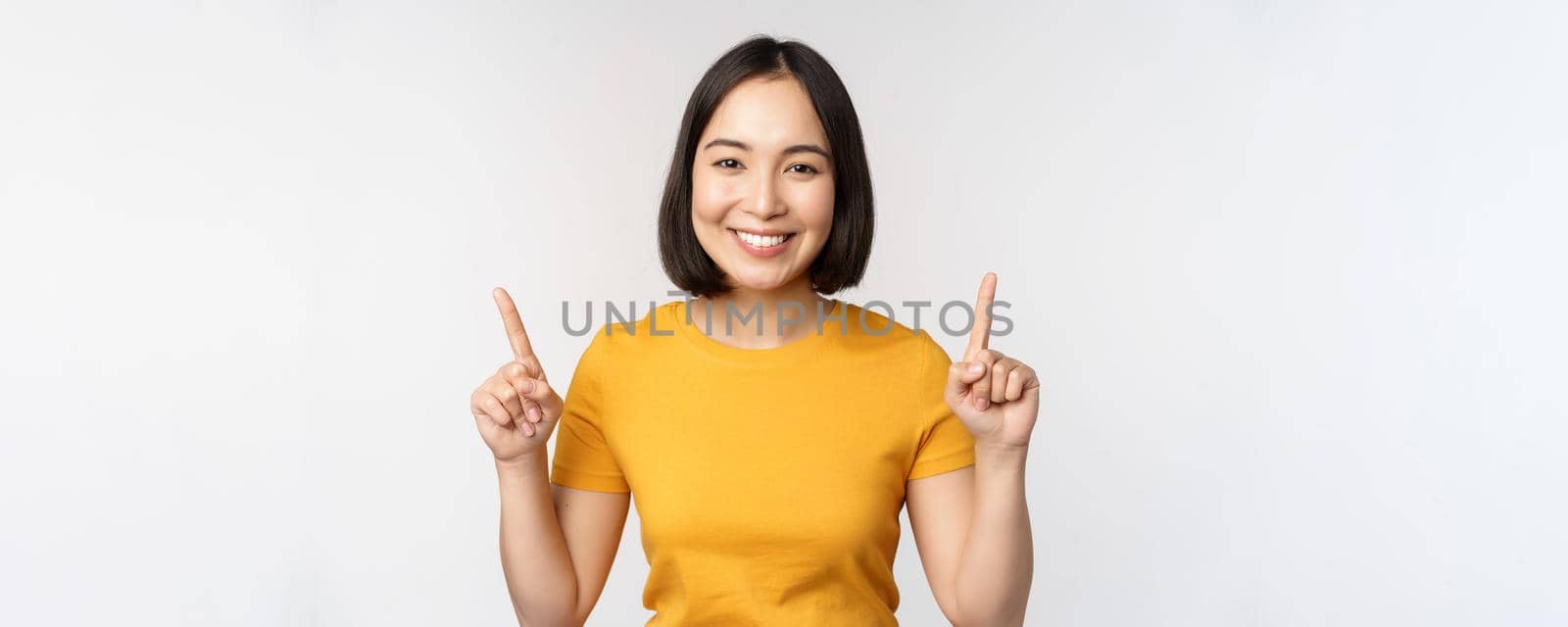 Portrait of beautiful japanese girl smiling, pointing fingers up, showing advertisement, standing in yellow tshirt against white background by Benzoix