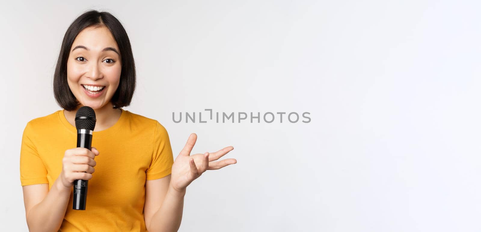 Image of young asian woman talking in microphone, perfom with mic, giving speech, standing in yellow tshirt against white background.
