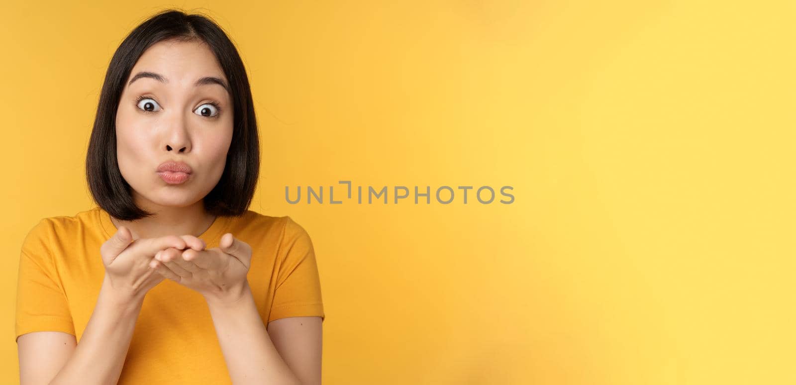Cute asian girl sending air kiss, blowing mwah with puckered lips, standing over yellow background by Benzoix