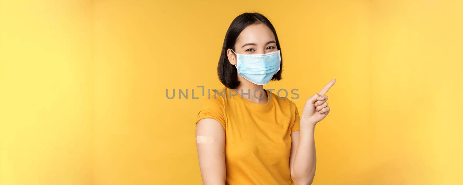 Vaccination from covid and health concept. Image of smiling korean girl in medical face mask, band aid on shoulder, pointing finger at banner advertisement, yellow background.
