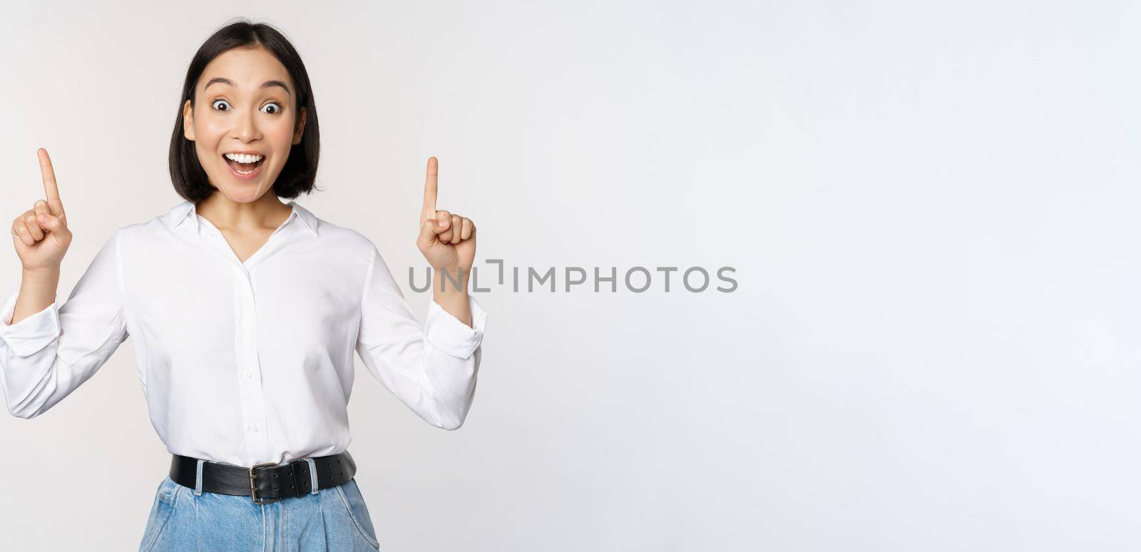 Enthusiastic business woman, asian female model pointing fingers up and smiling, making announcement, showing logo banner on top, white background.