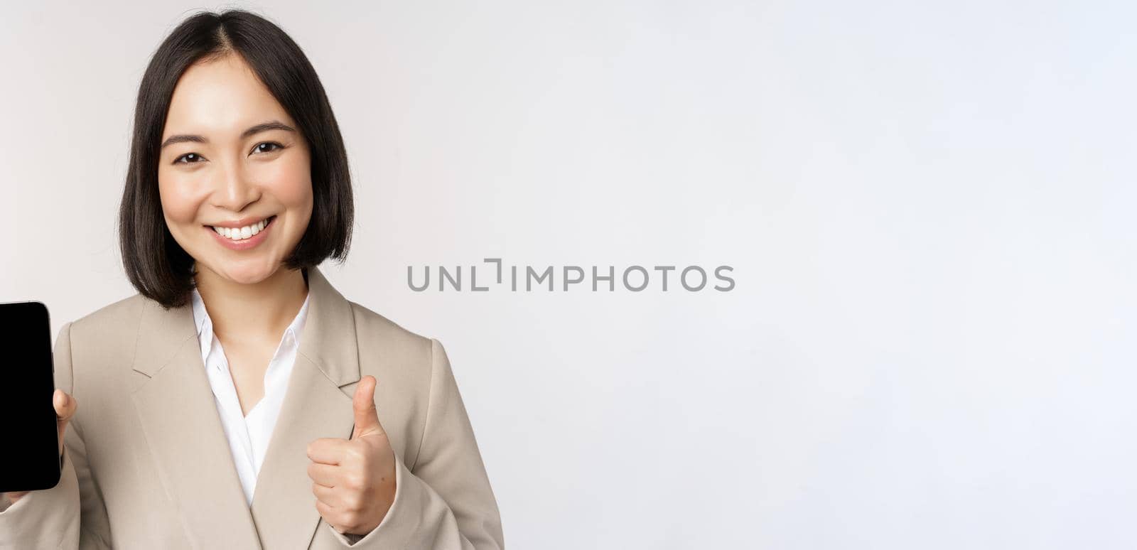 Smiling asian woman showing smartphone screen and thumbs up. Corporate person demonstrates mobile phone app interface, standing over white background.