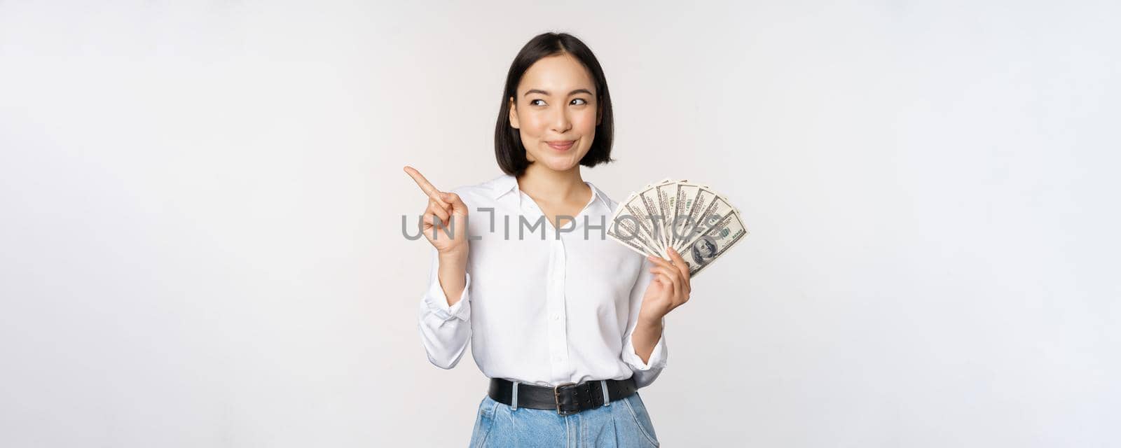 Smiling young modern asian woman, pointing at banner advertisement, holding cash money dollars, standing over white background by Benzoix