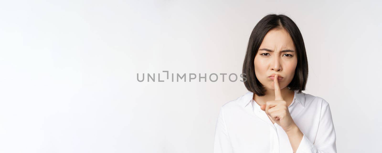 Close up portrait of young asian woman making hush, shhh shush sign, press finger to lips, dont speak, keep quiet gesture, white background by Benzoix