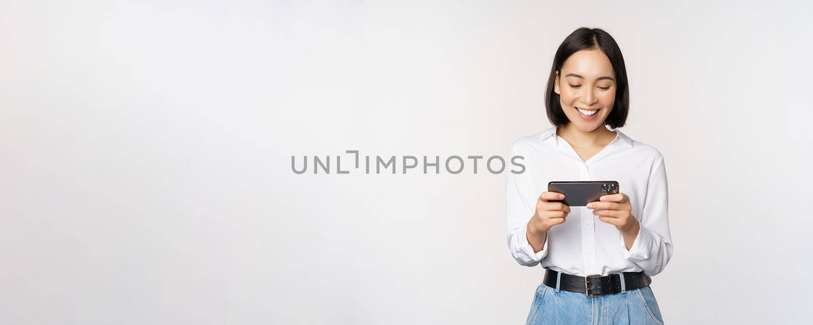 Young korean woman, asian girl playing mobile video game on smartphone, looking at horizontal phone screen, standing over white background by Benzoix