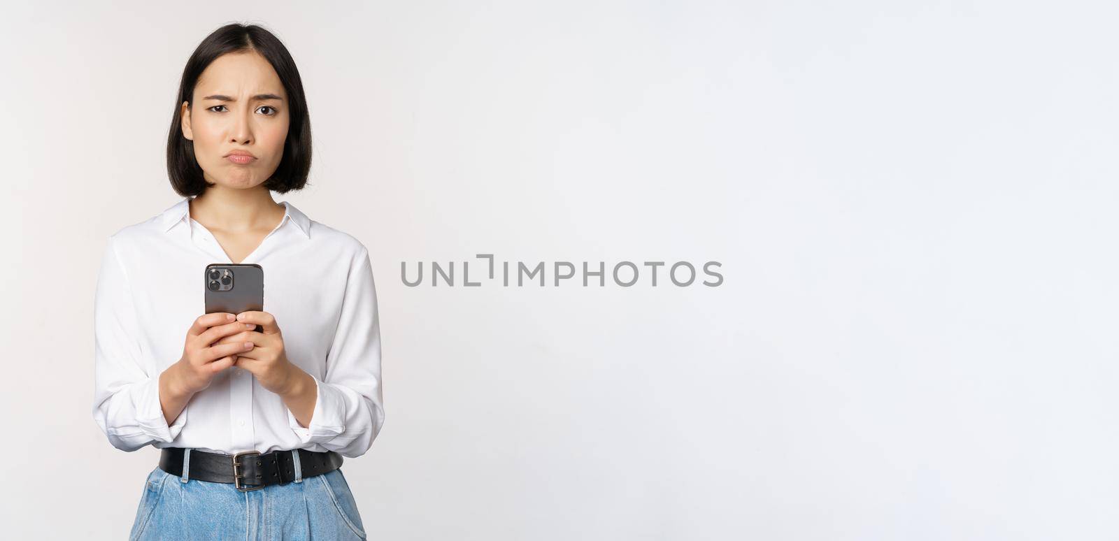 Asian woman holding smartphone and looking with doubt, disappointed with mobile phone app, standing agaist white background by Benzoix